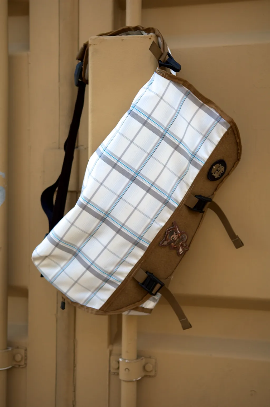 Kerberos bag hanging against a tan wooden crate wall, showing the plaid flap and shoulder strap